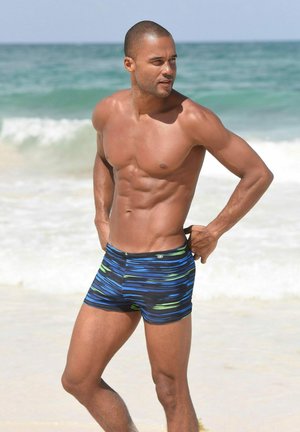 Blaue und schwarze gestreifte Badehosen mit grünen Akzenten, eng anliegend, aus dehnbarem Material, getragen von einer muskulösen Person am Strand.