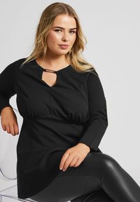 Musta pitkähihaisen tunika, jossa on avainaukko kaula-aukko ja muotoiltu vyötärö, yhdistettynä kiiltäviin mustiin legginsseihin. Suora kankaan rakenne.