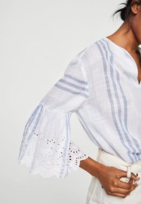 Blouse blanche légère à rayures horizontales bleues, col en V profond et manches évasées ornées de dentelle avec découpes œillets.