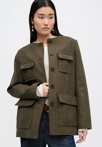 Brązowa kurtka w kratę z okrągłym dekoltem, czterema przednimi kieszeniami i dużymi czarnymi guzikami. Noszona na białej bluzce o fakturowanej powierzchni.