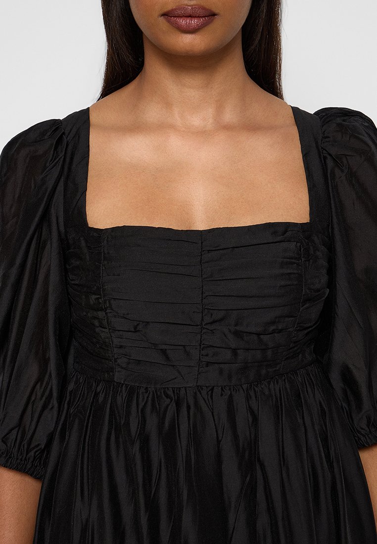Femme portant une robe noire avec un décolleté carré, un corsage froncé et des manches bouffantes arrivant au coude.