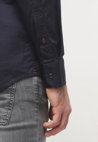 Tmavě modrá košile na knoflíky s kontrastními červenými a tmavě modrými knoflíky na manžetě, vyrobená z hladké, lehké tkaniny a střih přizpůsobený postavě.