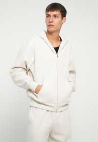 Felpa con zip di un beige chiaro, realizzata in tessuto morbido. Presenta una tasca frontale, cappuccio regolabile e vestibilità relaxed con polsini a coste.
