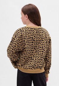 Niña con cabello largo y castaño, vistiendo un suéter de estampado de leopardo en tonos marrón claro y negro, y pantalones negros, de espaldas contra un fondo claro y sencillo.