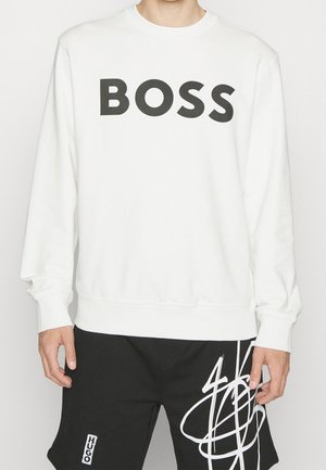 Mężczyzna ubrany w biały sweter z napisem "BOSS" dużymi czarnymi literami oraz czarne szorty z białym abstrakcyjnym wzorem i napisem "HUGO".