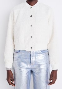White textured cropped jacket with a round collar, featuring metal buttons and knit cuffs. Paired with shiny light blue metallic pants.