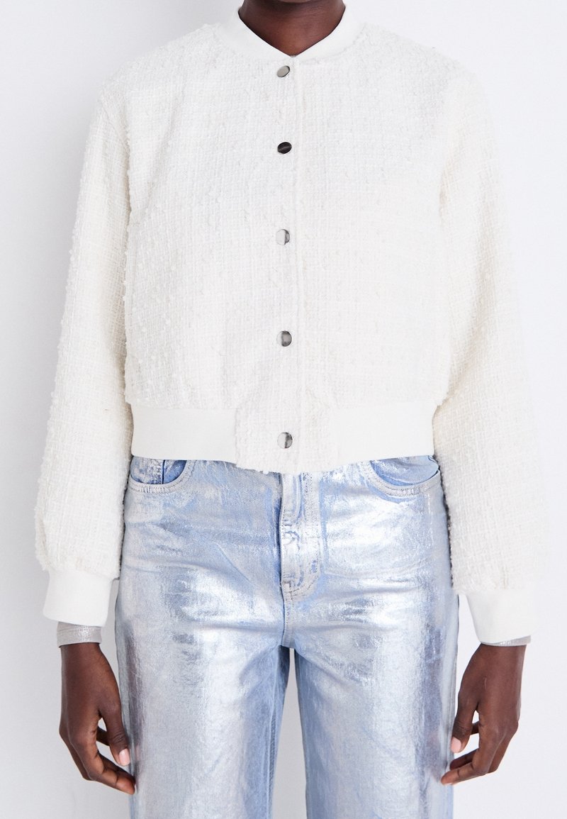 White textured cropped jacket with a round collar, featuring metal buttons and knit cuffs. Paired with shiny light blue metallic pants.