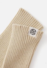 Gants sans doigts en tricot côtelé beige avec une finition texturée et une petite étiquette blanche cousue portant un logo noir.