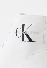 Weißer Canvas-Sneaker mit einem auffälligen schwarzen "CK"-Logo und dem Schriftzug "CALVIN KLEIN JEANS". Strukturierte Oberfläche mit Metallösen für die Schnürsenkel.