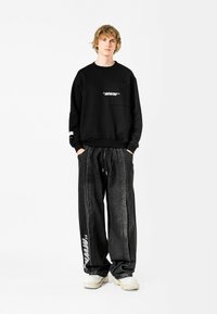 Sudadera negra con un bolsillo frontal y texto "MVM" en blanco; combinada con pantalones de denim oscuros y sueltos que tienen un logo en el costado. Zapatos amarillentos.