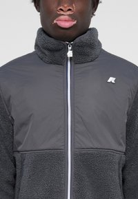 Veste grise en matériaux mixtes : partie supérieure lisse et partie inférieure en polaire texturée, col haut, fermeture éclair à l'avant et accent logo.