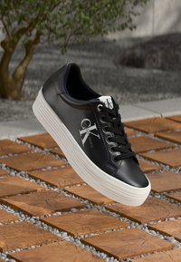 Zapatilla de cuero negro con suela blanca, cordones y logotipo. Presenta una punta redondeada y una superficie texturada. Se eleva sobre una superficie con patrón de ladrillo.