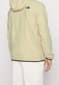 Beige huva jacka med elastiska ärmslut och svart fåll. Har The North Face-logotypen på baksidan. Mjuk, texturerad tyg.