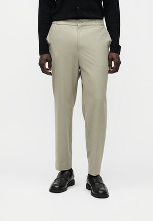 Mand iført beige skræddersyede bukser, sort langærmet skjorte og sorte læderloafers, stående på en hvid baggrund.