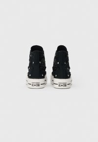 Must be musta konversi All Star mustam kõrge tippu kingad, millel on hõbedased nupud ja mustad kangakimbud, nähtavad tagantpoolt valgel taustal.