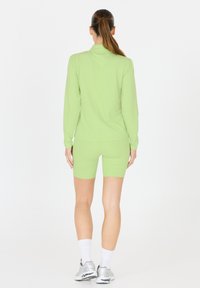 Femme avec une queue de cheval brune portant un haut à manches longues vert clair et un short assorti, des chaussettes blanches et des baskets argentées, debout de dos.