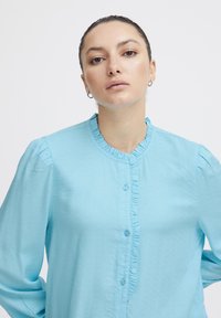 Blusa button-up azzurro chiaro realizzata in tessuto testurizzato. Presenta un colletto e maniche arricciate, con bottoni coordinati lungo la parte anteriore.