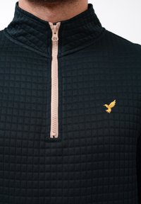 Svart kviltet hettegenser med glidelås, med en beige glidelås som har brodert en liten gullfugl på venstre bryst. Myk tekstur og tettsittende design.