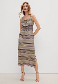 Maxi-Kleid mit einem Zickzackmuster in lila, gelben und grünen Farbtönen, mit tailliertem Bund und seitlichem Schlitz. Kombiniert mit bunt gemusterten Sandalen.