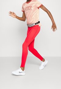 Person som springer iförd en rosa Nike-t-shirt, ljusröda leggings, vita Nike-sneakers och en mönstrad midjeväska.