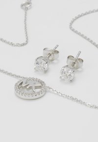 Conjunto de joyería de plata que presenta pendientes redondos con piedras transparentes y un collar tipo pendant con un diseño circular y logotipo grabado.