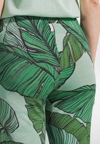 Grüne Leggings mit einem großen, blattartigen Muster in verschiedenen Grüntönen und schwarzen Konturen. Weicher Stoff, figurbetontes Design, taillenhoher Schnitt.