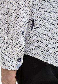 Chemise blanche à pois navy, à manches longues, avec détails de boutons et tissu contrastant au niveau des poignets. Étiquette de la marque visible près de l'ourlet.