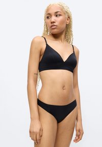 Jeune femme aux longues tresses blondes portant un bikini noir, debout devant un fond clair uni, regardant sur le côté.