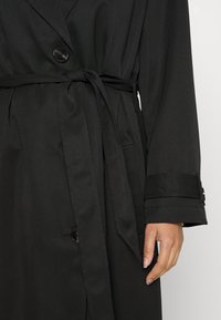 Svart trenchcoat med belte i midjen, lange ermer og enkelt knappelukking. Laget av glatt stoff med en matt finish.