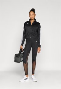 Črna atletska jakna z belimi črtami, v kombinaciji s črnimi legicami s prozornimi paneli. Vključeni so beli supergi in črna športna torba.