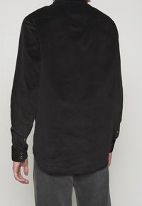 Camicia nera in velluto a coste con maniche lunghe, polsini con bottoni e orlo arrotondato. Presenta coste verticali e dettagli di cucitura discreti sulle spalle.