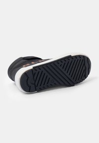 Schwarzer Sportschuh mit einer strukturierten Gummisohle, ausgestattet mit einem Klettverschluss und einer kontrastierenden weißen Zwischensohle. Einfaches Design mit minimalen Akzenten.