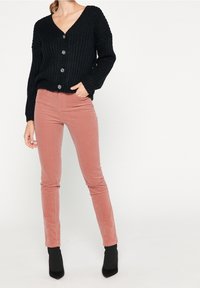 Svart stickad cardigan med djup V-ringning, fem knappar och ribbad textur, matchad med rosa kordflöjt slim-fit byxor och svarta ankelskor.