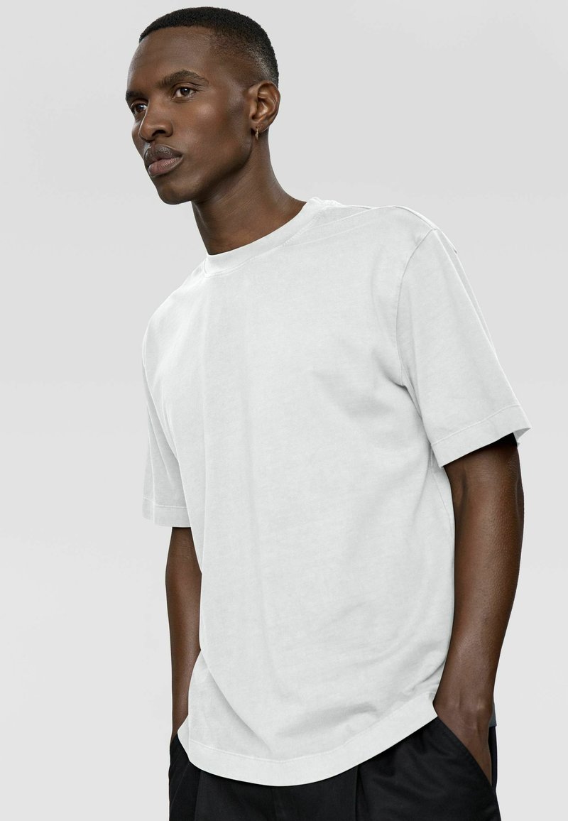 Jeune homme aux cheveux courts et portant une boucle d'oreille, vêtu d'un t-shirt blanc à manches courtes uni et d'un pantalon noir, debout avec les mains dans les poches.