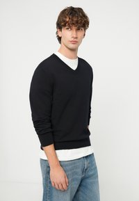 Młody mężczyzna w czarnym swetrze z dekoltem w serek na białej koszulce i niebieskich dżinsach, patrzący lekko na bok na tle jednolitej białej ściany.