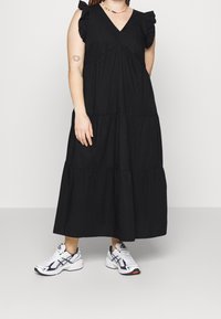 Femme portant une robe maxi noire sans manches à étages avec des épaules froufroutées et des baskets blanches, debout devant un fond clair uni.