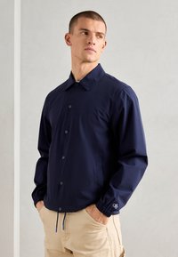 Navy lightweight jacket with snap buttons, elastic cuffs, and two side pockets. Features a subtle logo accent on the sleeve.