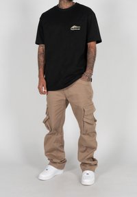 Svart bomulls-T-shirt med liten logga på bröstet, kombinerad med lösa beige cargobyxor med flera sidofickor och vita sneakers.