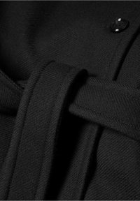 Primo piano di un tessuto nero con una trama texturizzata, un dettaglio della cintura e un accento di bottone, che enfatizzano un aspetto liscio e sartoriale.