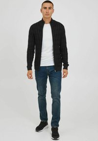 Black ribbed cardigan with a zip closure, worn over a white shirt. Paired with blue jeans and black sneakers. Simple and fitted design.