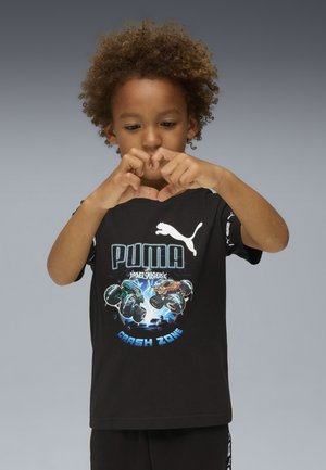 Bambino con capelli ricci che indossa una maglietta nera Puma "Crash Zone" mentre forma un cuore con le dita su uno sfondo grigio.