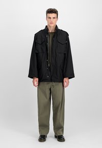 Homme debout devant un fond uni, portant une veste noire, un pantalon vert olive et des chaussures noires, fait face à l'avant, les bras détendus.