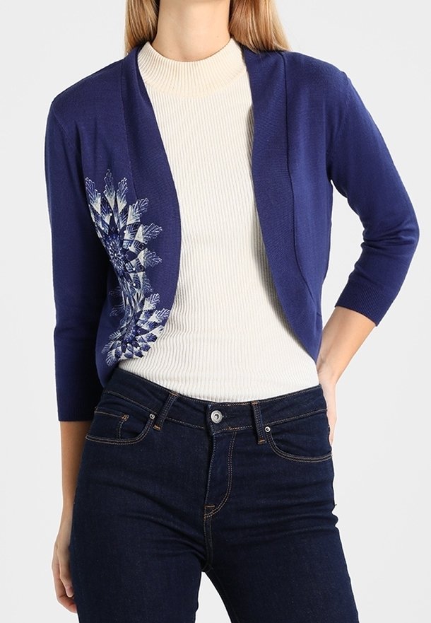 Femme portant un gilet court bleu marine avec une broderie florale blanche et bleue sur un col roulé blanc côtelé et un jean foncé.