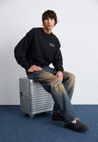 Sudadera negra con logo, vaqueros de denim azul deslavado con rotos y zapatos de plataforma negros, sentado en una maleta plateada y estriada.