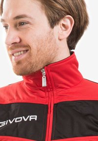 Giacca rossa e nera con colletto alto, chiusura con zip e dettaglio del logo. Realizzata in tessuto liscio con accenti e cuciture a contrasto.