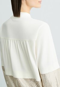 Lightweight cream top with a pleated back and shimmering metallic gold fabric at the lower hem. Smooth texture and relaxed fit.