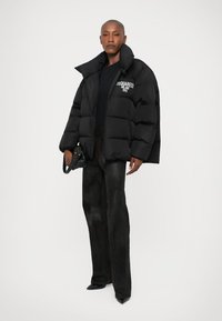 Zwarte pufferjack met een hoge kraag, voorzien van een witte logo op de linkerkant van de borst. Gestyled met donkere broek en elegante zwarte schoenen.