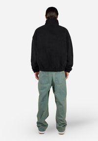 Persona con cabello corto y oscuro, vistiendo una chaqueta de forro polar negra, unos vaqueros sueltos de color azul claro y zapatillas blancas, de pie de espaldas sobre un fondo blanco.