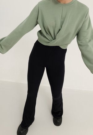 Personne portant un haut à manches longues vert sauge torsadé à la taille, un pantalon noir ajusté évasé, et des chaussures noires épaisses sur fond clair.