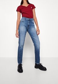 Red v-neck t-shirt, blue jeans with a slim fit, and black chunky shoes. Denim texture is visible; shirt features a small logo left side.